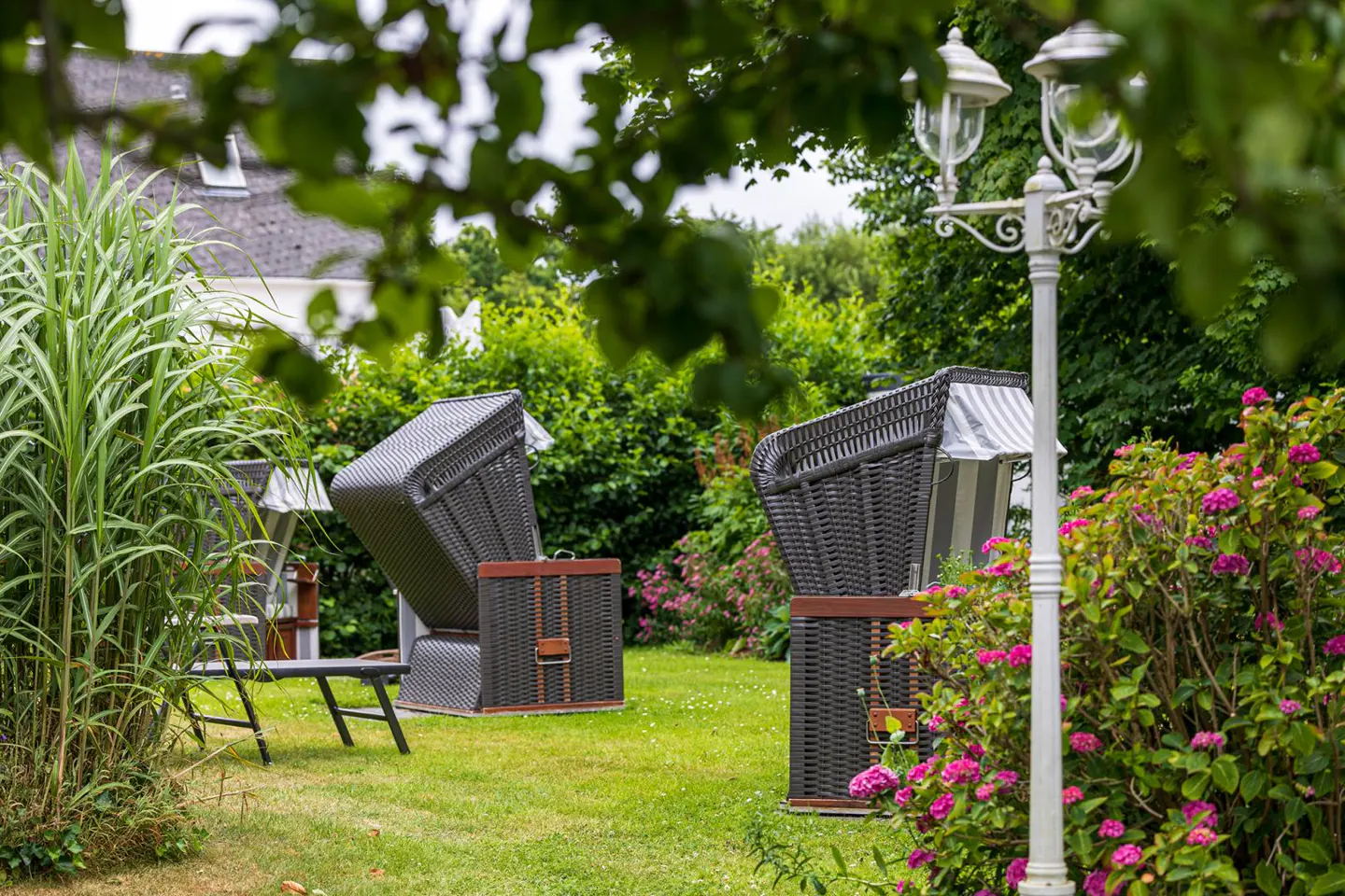 Lush green lawn with two dark wicker beach chairs, pink flowers, and a white lamp post. Tall grass and trees in the background.