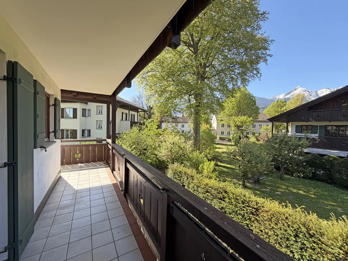 Balcony view with tile floor, dark wood railing, and green shutters. A lawn, trees, buildings, and snow-capped mountains are visible.