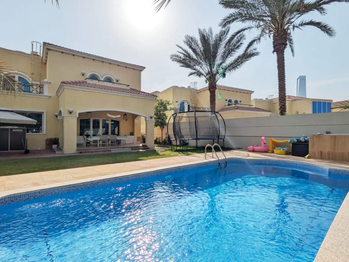 Backyard view of a tan two-story house with a blue swimming pool, palm trees, and a trampoline.