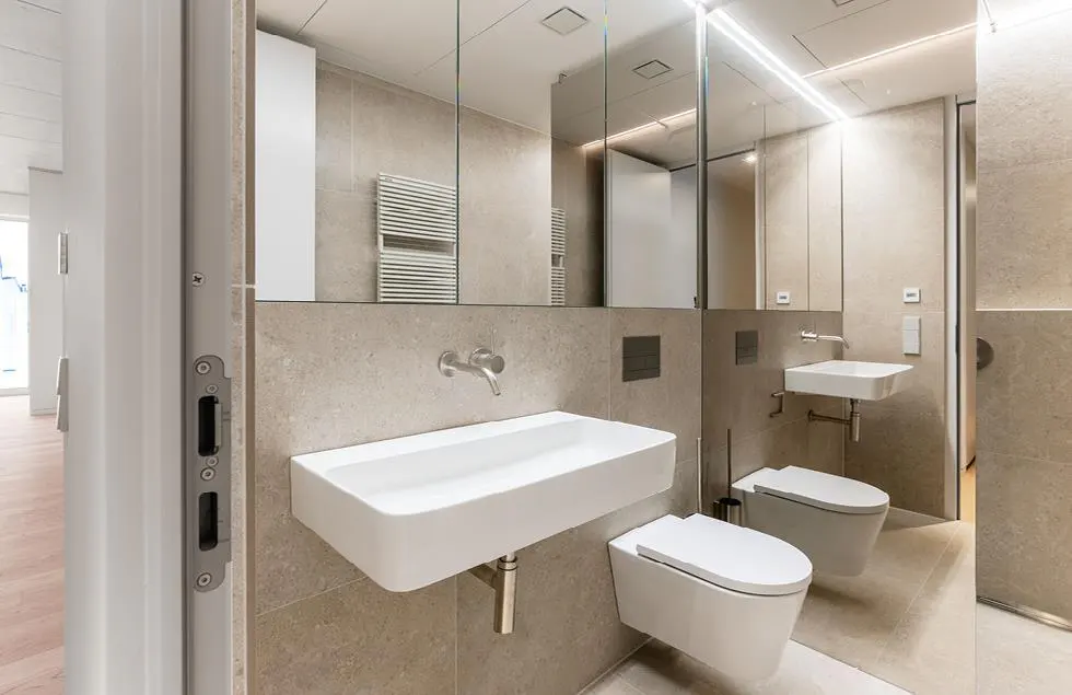 Modern bathroom with beige tile walls, white rectangular sinks and toilets. A large mirror reflects the room. Open door on the left.