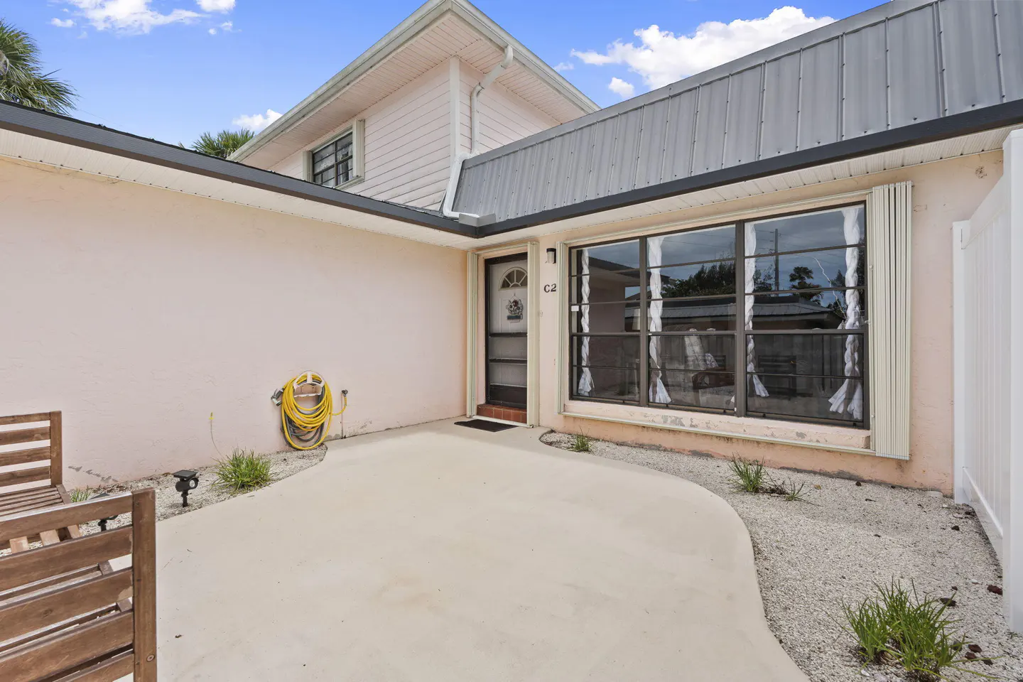 Exterior view of a pink house with a gray metal roof, a black door, and a large window. A yellow hose is coiled on the wall. A wooden bench sits on the patio.