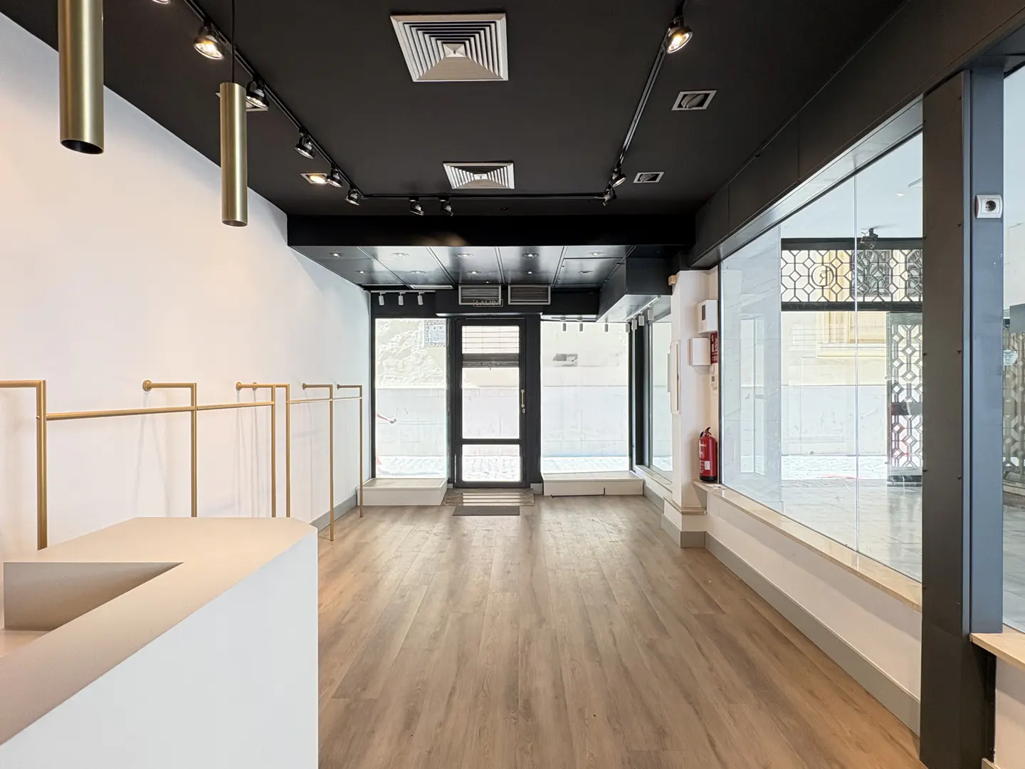 Empty retail space with wood floors, white walls, and a black ceiling with track lighting. Clothing racks line the left wall. Large windows face the street.