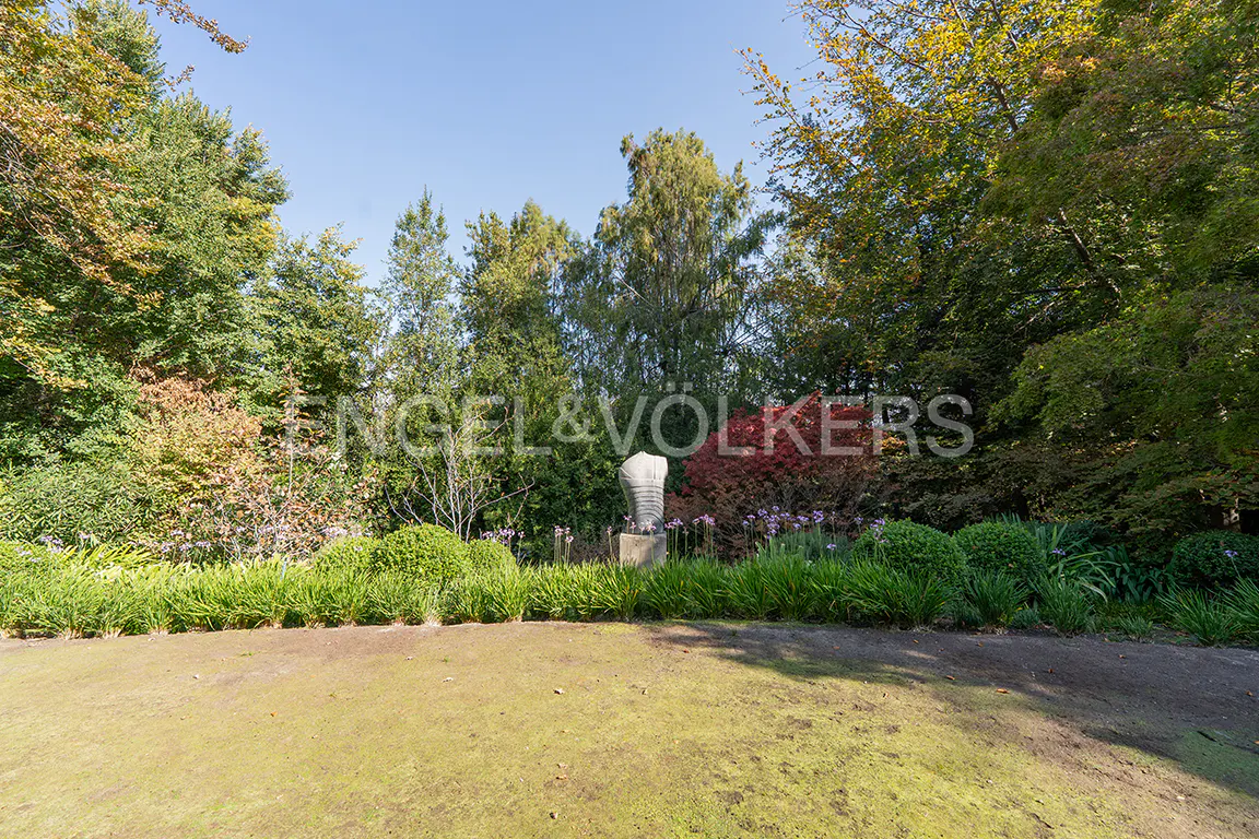 Lush green lawn and trees surround a modern sculpture in a garden setting under a clear blue sky.