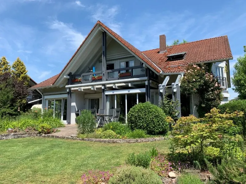 Two-story house with a red tile roof, gray trim, and a green lawn with flowers and bushes. A patio with a table and chairs is visible.