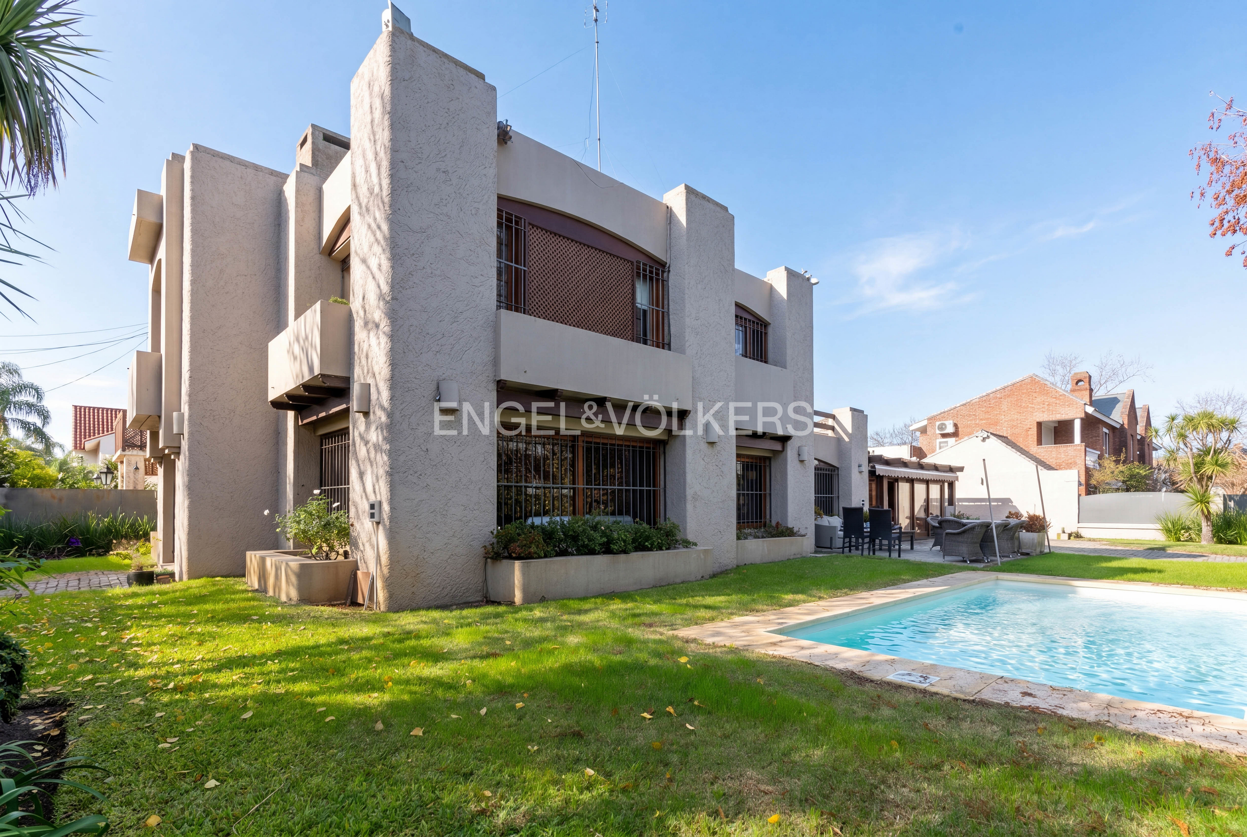 Modern beige two-story house with a swimming pool, green lawn, and patio furniture under a clear blue sky.