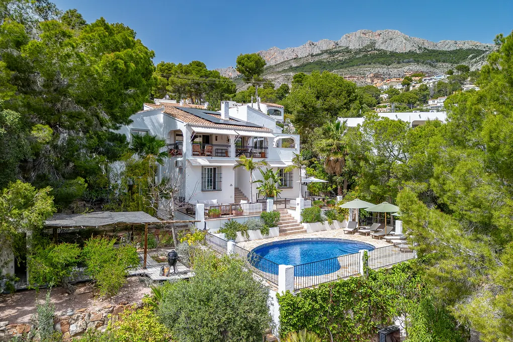 Two-story white house with a blue pool, surrounded by green trees and a mountain in the background.