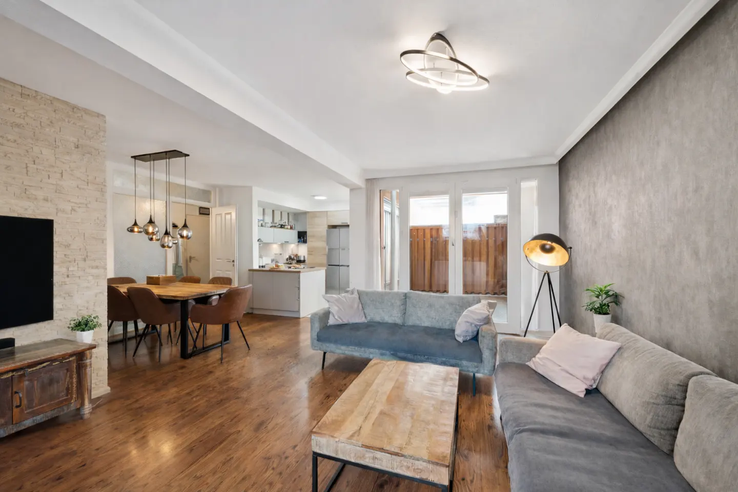 Open-concept living space with wood floors, gray sofas, and a dining area with a wooden table and brown chairs. A kitchen is visible in the background.