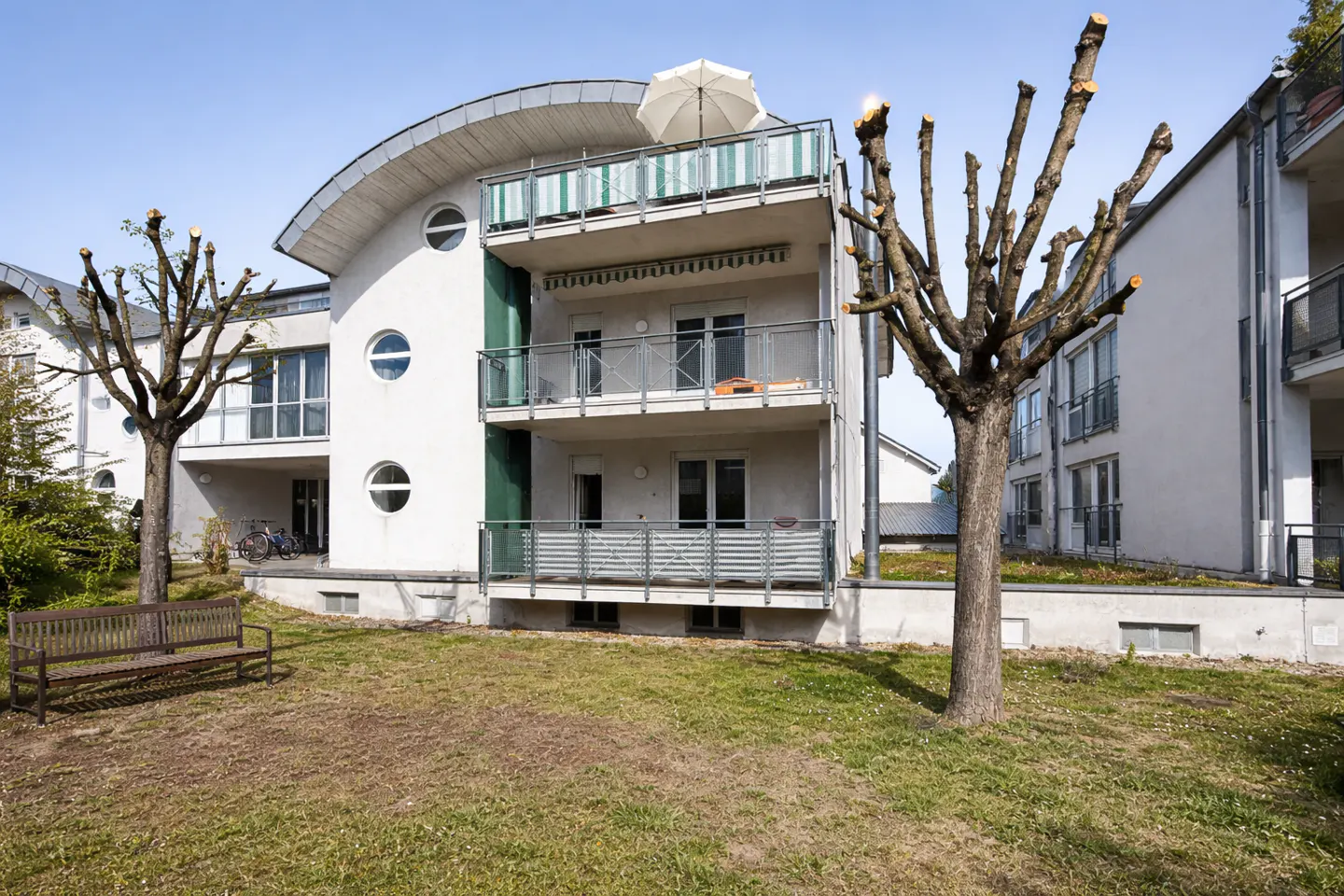 Modern white apartment building with balconies and round windows, set on a green lawn with a wooden bench and pruned trees.