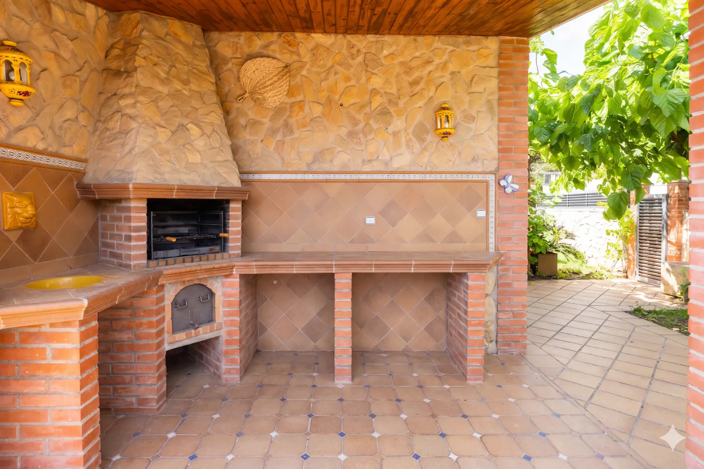 Outdoor kitchen with brick oven, stone wall, and tiled floor. A yellow lantern hangs on the wall. Green trees are visible outside.