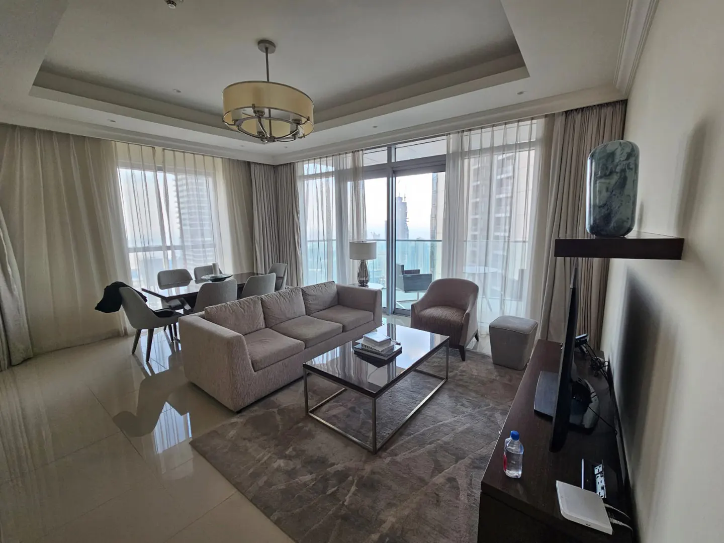 A modern living room with a gray sofa, glass coffee table, and dining set. Floor-to-ceiling windows offer a city view. A TV and decor sit on a dark wood console.