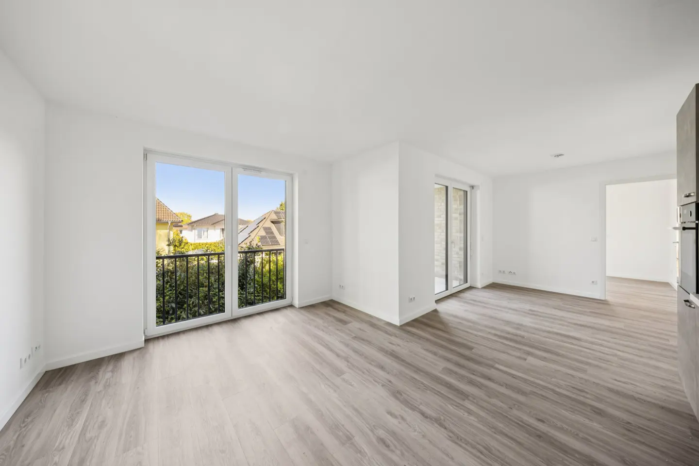 Bright, empty room with white walls and gray wood floors. Large windows lead to a balcony with green foliage. A sliding glass door is on the right.