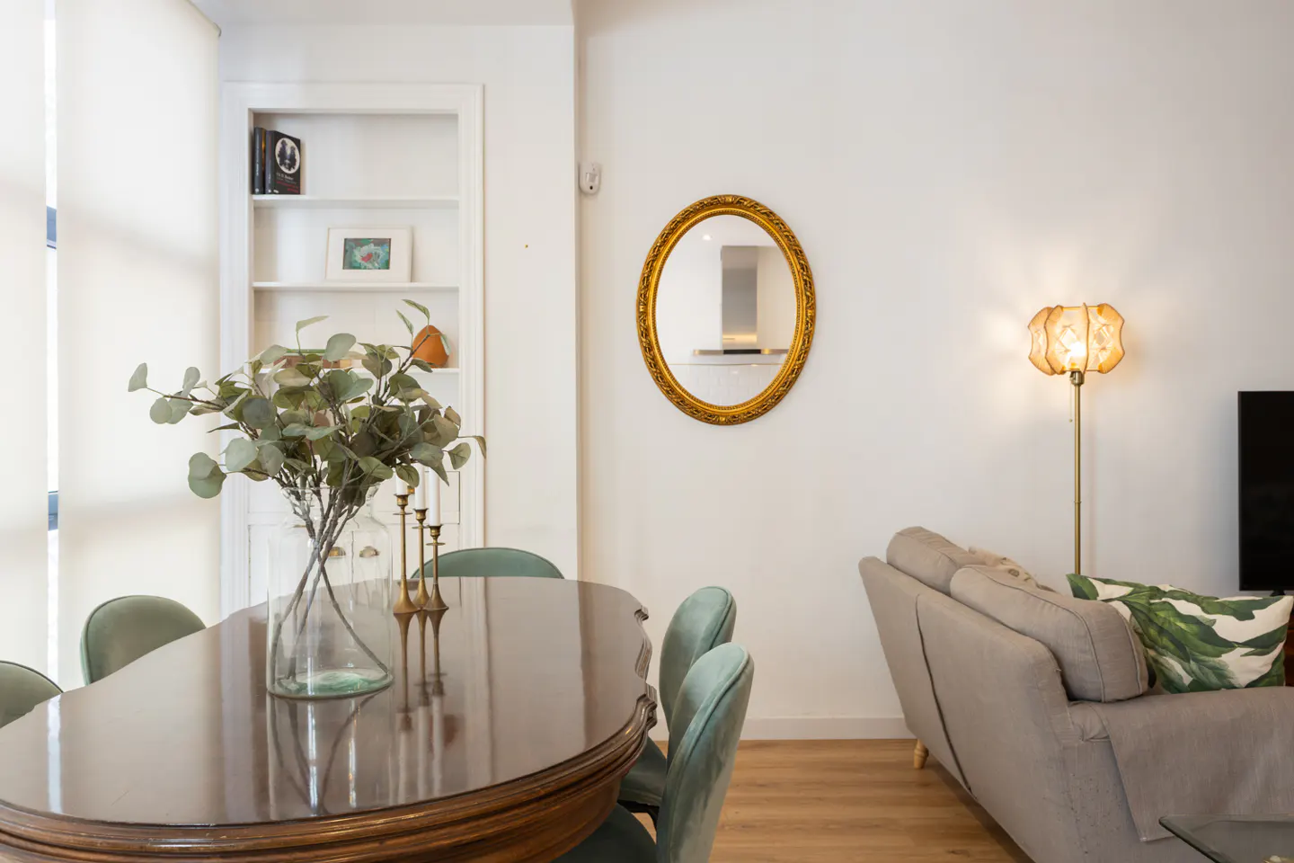 A bright living room with a dark wood table, green chairs, and a beige sofa. A gold mirror hangs on the white wall.