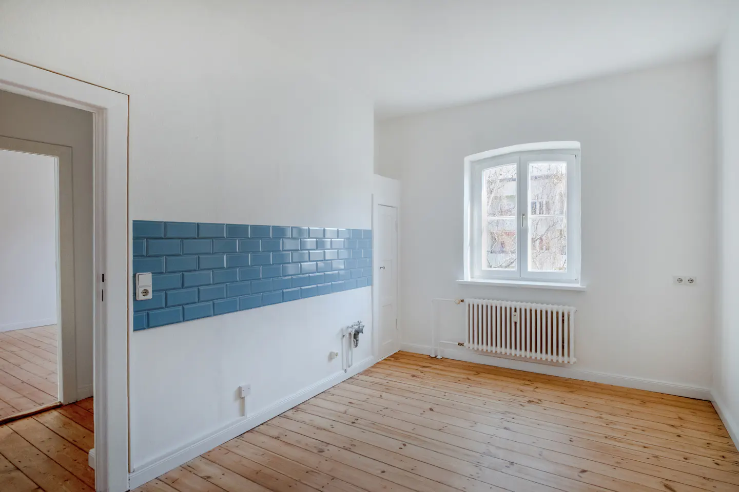 Bright, empty room with wood floors, white walls, blue tile accent, window, and radiator.