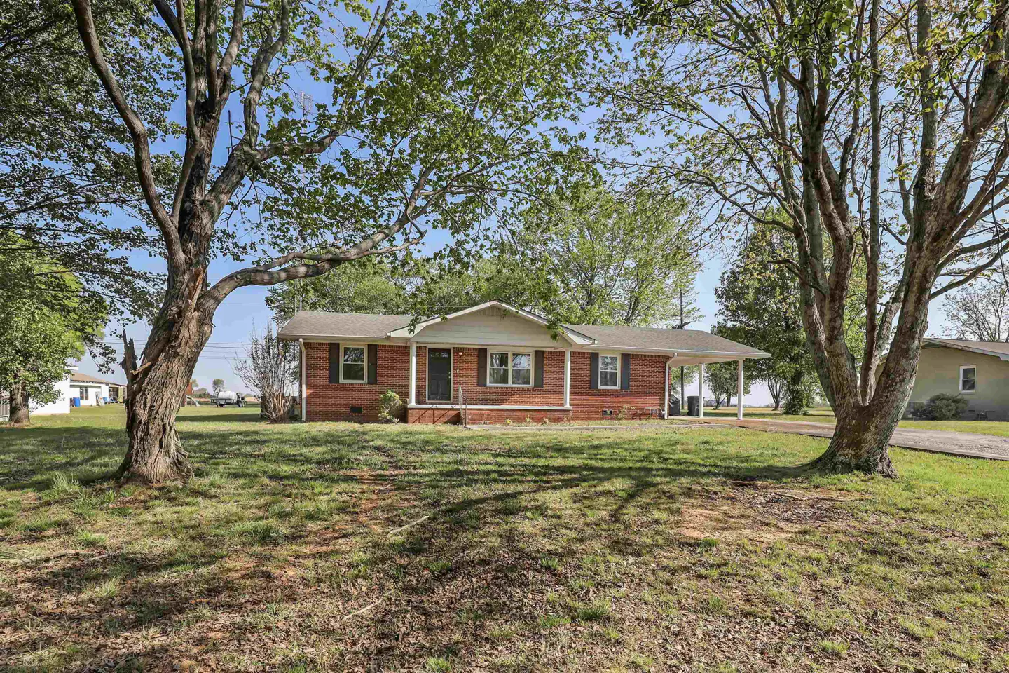A single-story red brick house with green shutters, a carport, and a large front yard with trees.