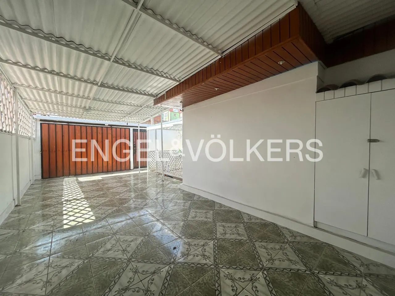 Covered patio with tile floor, white walls, and a corrugated metal roof. A brown wooden gate is visible in the background.