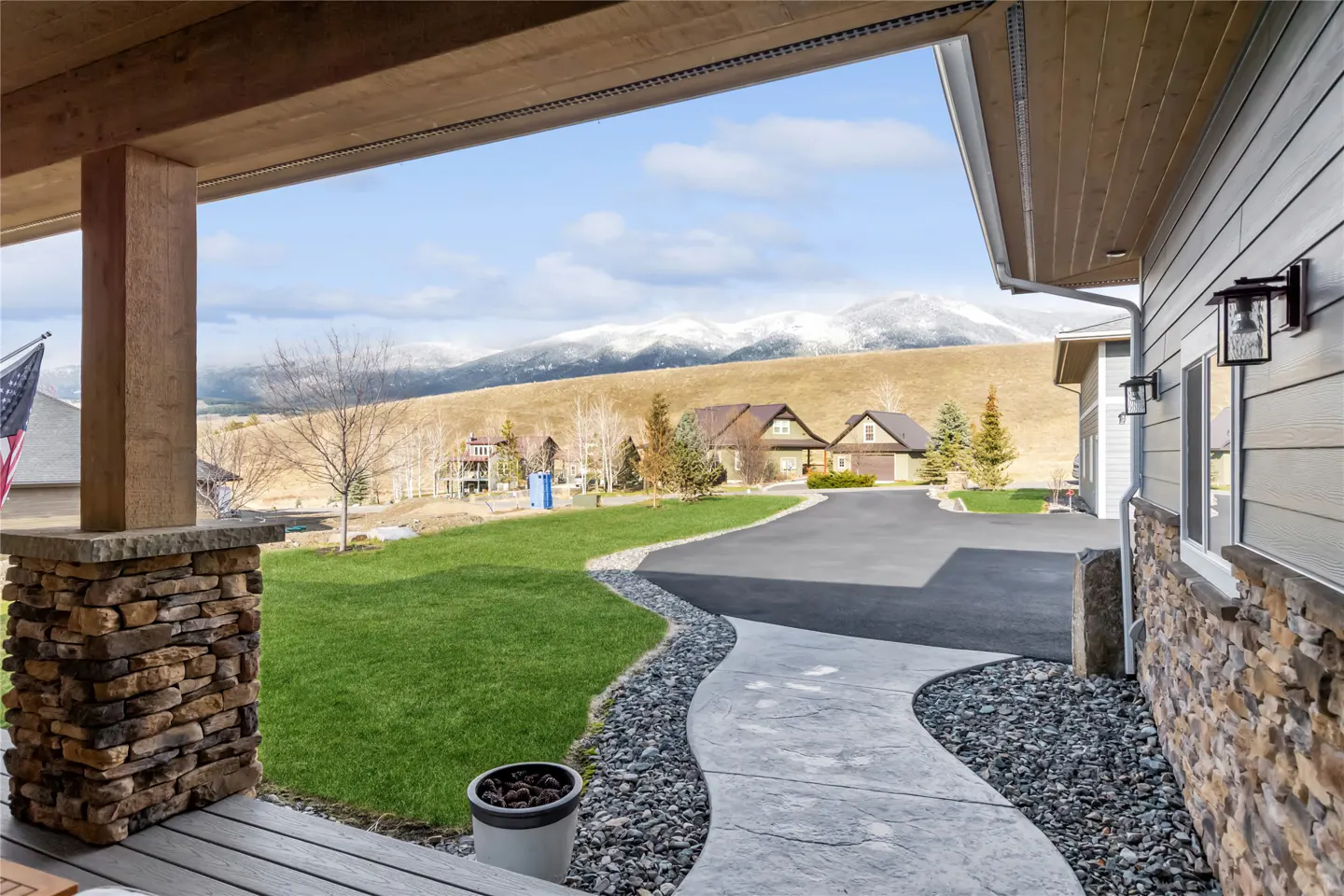 View from a porch with stone columns, looking out to a green lawn, driveway, houses, and snow-capped mountains.
