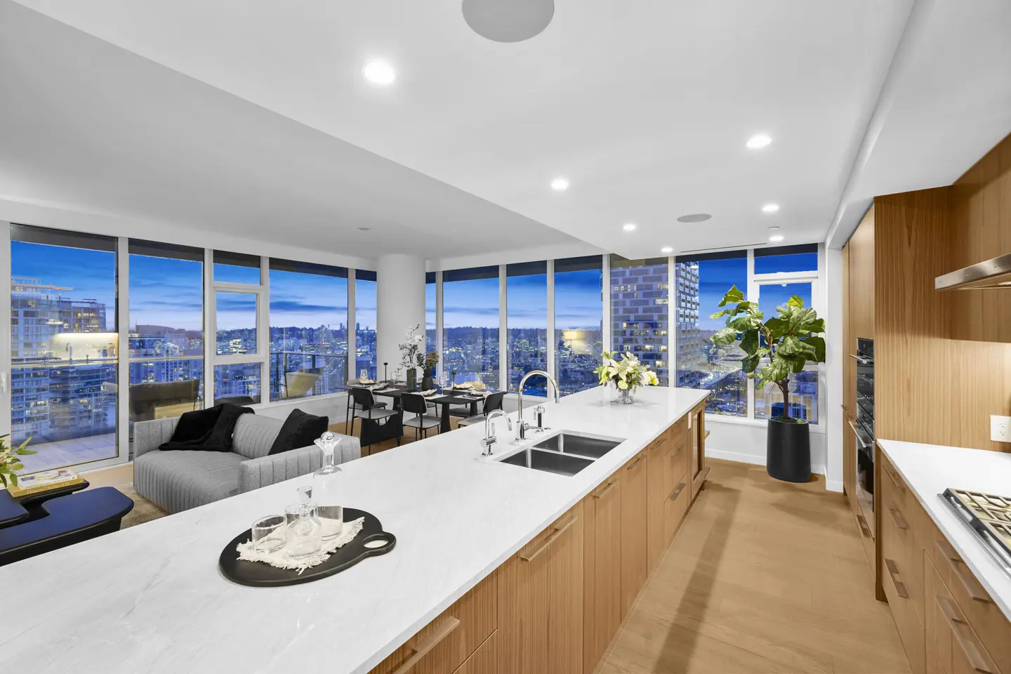 Bright, modern condo interior with a white countertop island, wood cabinets, and city views through large windows. A dining table and sofa are visible.