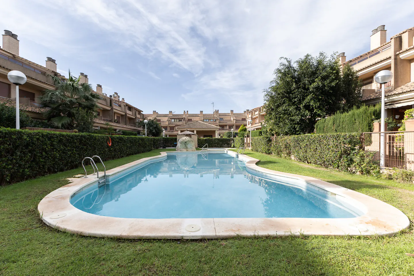 Outdoor pool with clear blue water, surrounded by green grass and trimmed hedges, with residential buildings in the background.