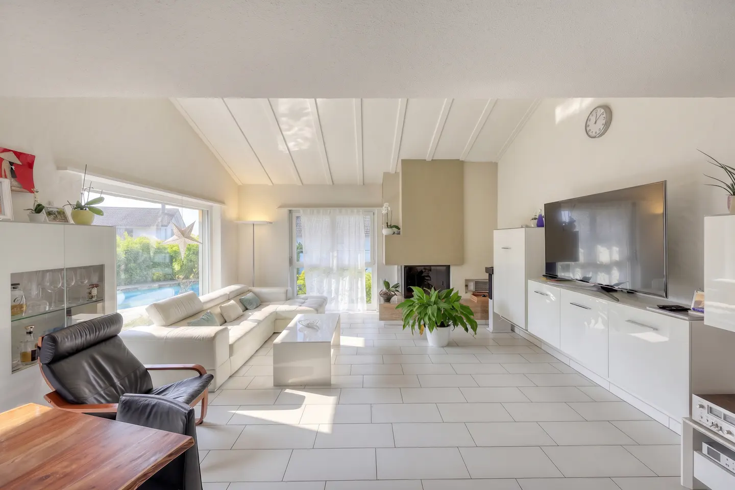 Bright living room with white tile floor, white sectional sofa, fireplace, and large TV. A black leather chair sits near a wooden table.