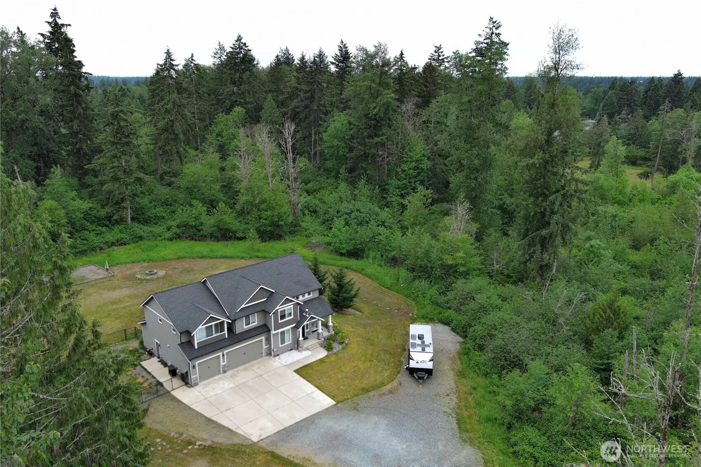 Aerial view of a gray two-story house with a dark roof, a concrete driveway, and a forest backdrop.