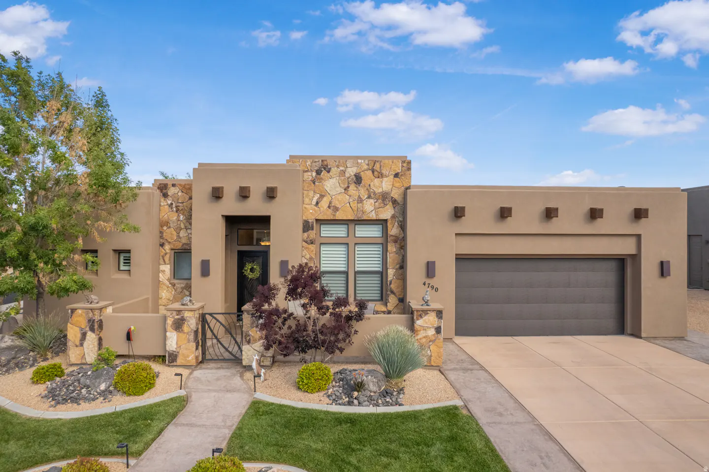 Tan stucco house with stone accents, a gray garage door, and a landscaped yard under a blue, cloudy sky.