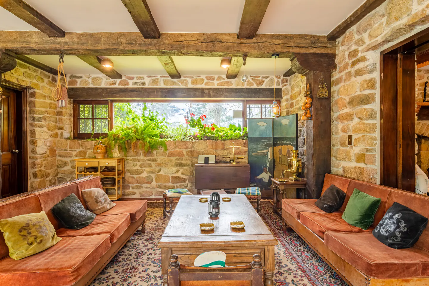 Living room with stone walls, exposed wood beams, and two rust-colored sofas facing each other. A large wooden coffee table sits on a patterned rug.