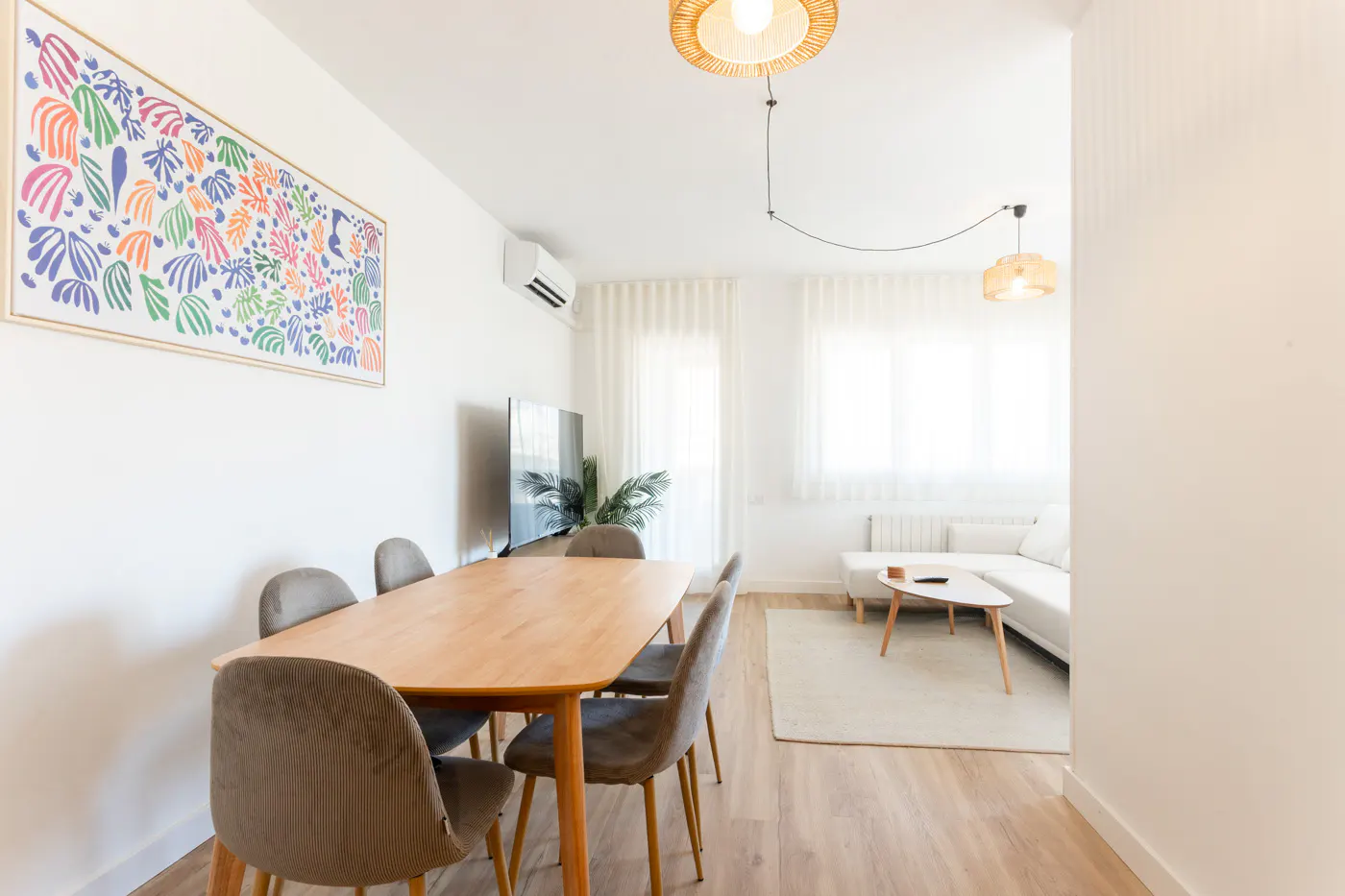 Bright living and dining area with a wooden table, grey chairs, white sofa, and colorful abstract wall art.