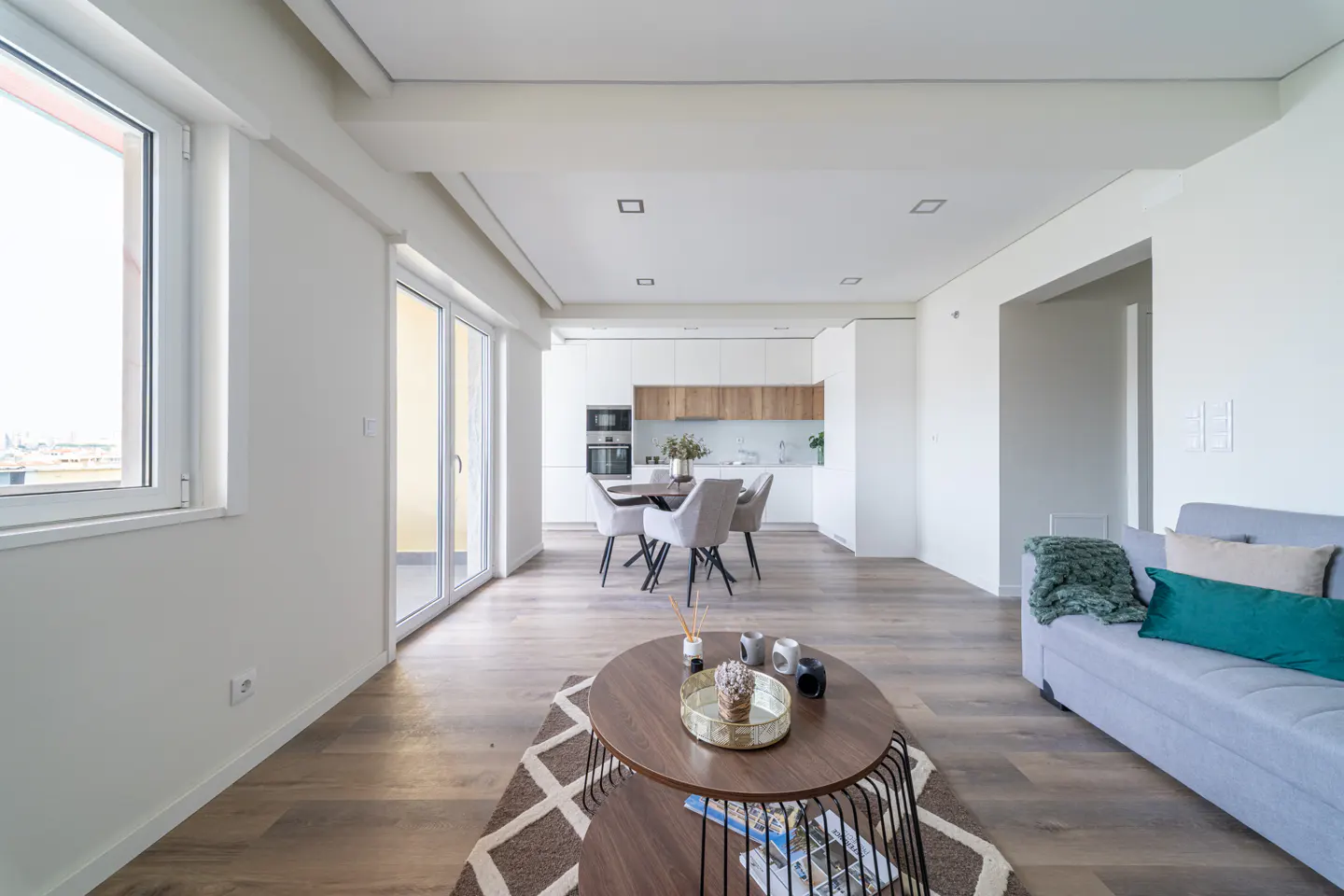 Bright, modern apartment interior with wood floors, white walls, and open kitchen. Gray sofa, oval coffee table, and dining set visible.