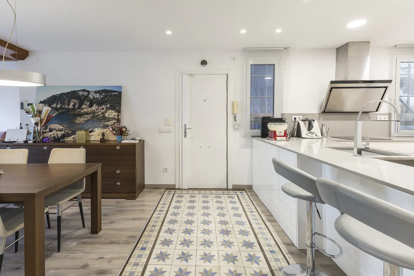 Bright, modern kitchen and dining area with white cabinets, stainless steel appliances, and a dark wood dining table. A patterned tile runner leads to the front door.