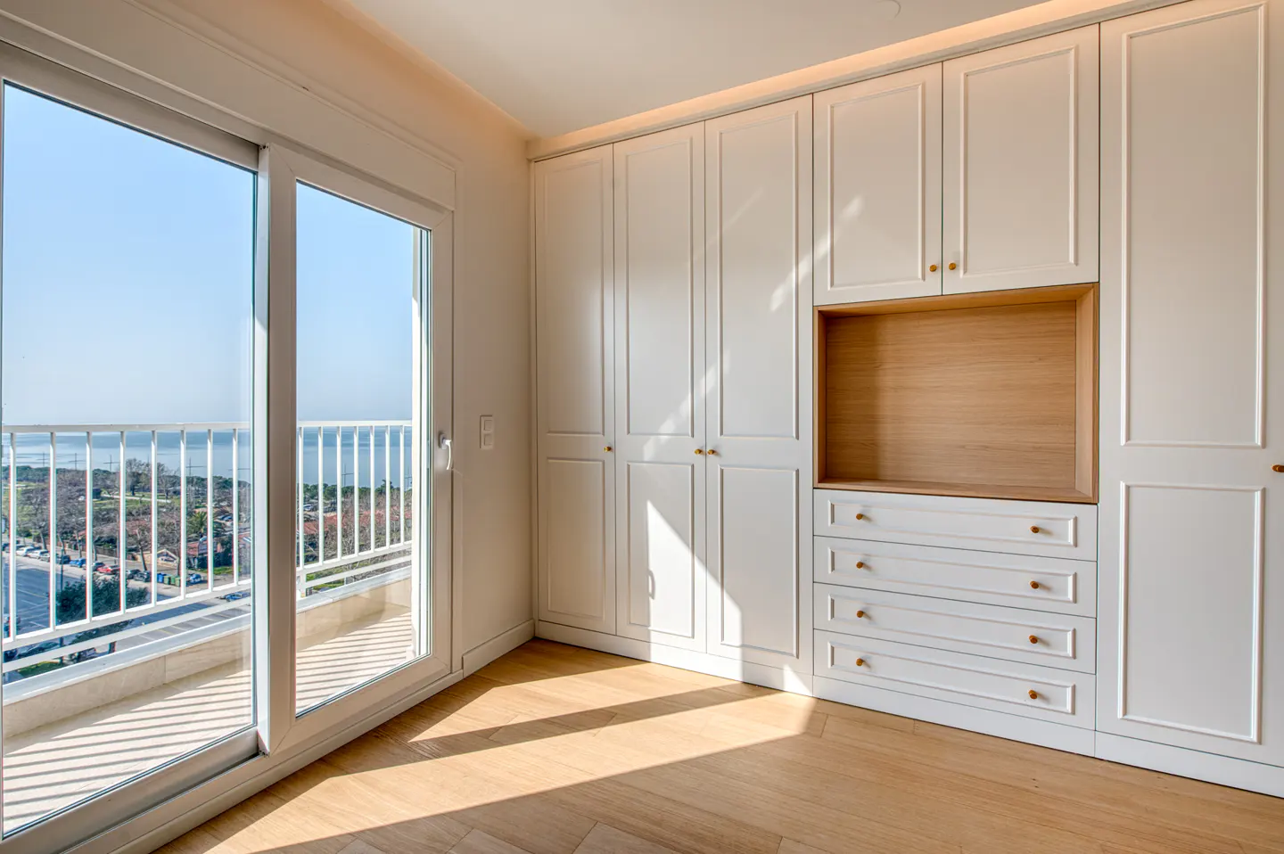 Bright bedroom with white built-in wardrobe, drawers, and a sliding glass door to a balcony with ocean view.