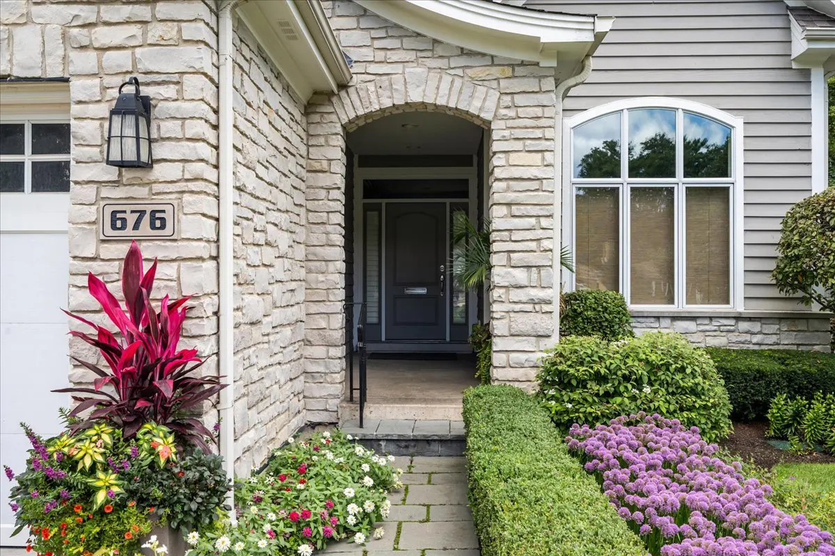 Stone house exterior with gray door, arched entrance, and address number 676. Flower beds and a stone walkway lead to the entrance.