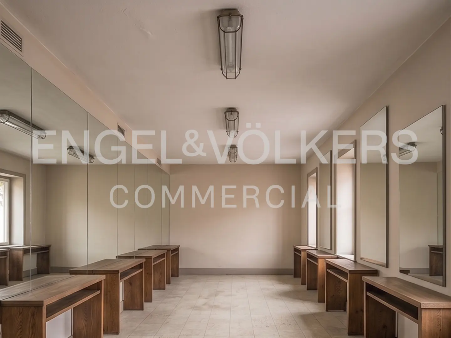 A bright, minimalist room with beige walls, tiled floors, wall-to-wall mirrors, and wooden vanity desks along both sides.