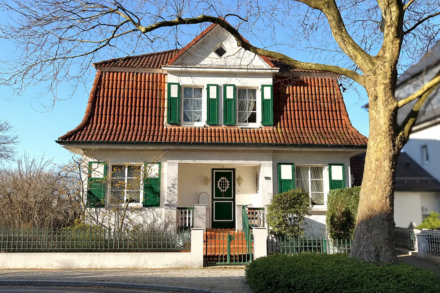 Two-story house with red tile roof, white walls, and green shutters. A tree frames the right side of the image.