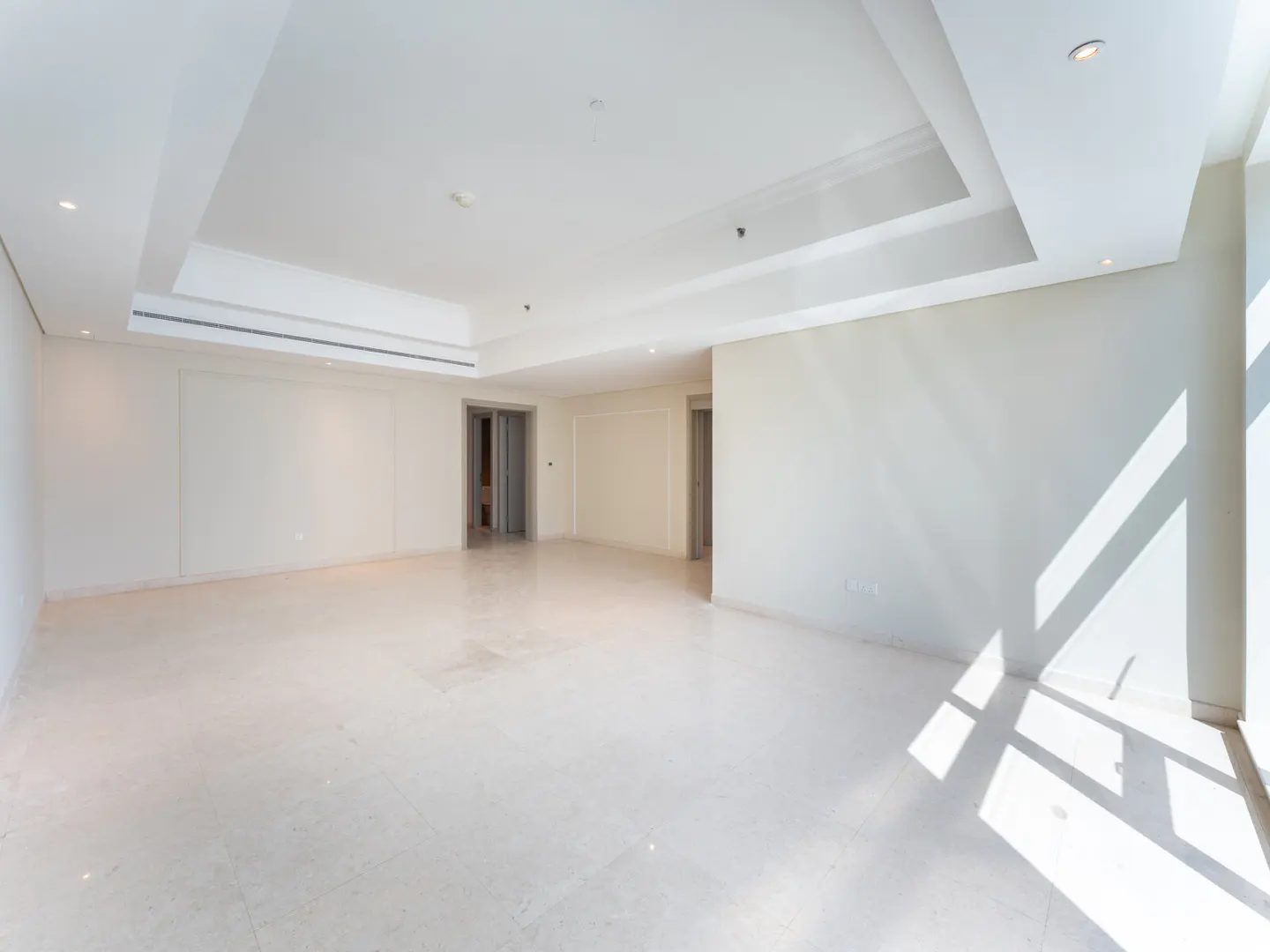 Bright, empty room with white walls, marble floor, and recessed lighting. Sunlight streams in from the right.