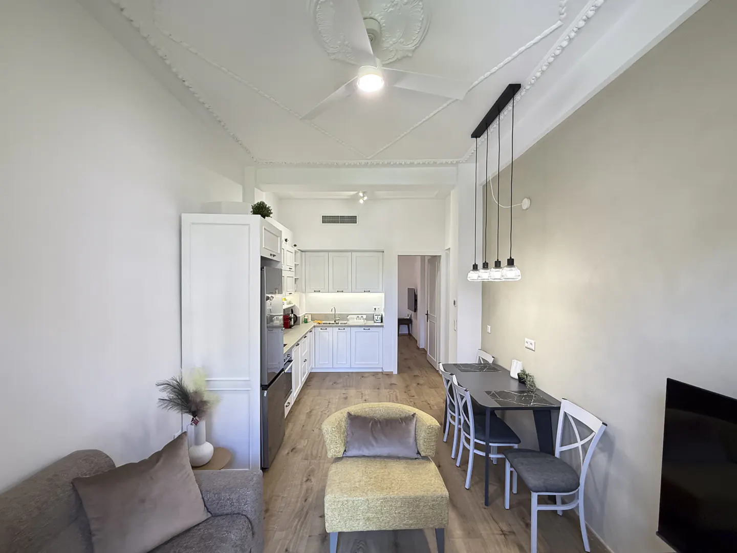 A bright, modern apartment with white walls, a kitchen, dining area, and living room. A ceiling fan and pendant lights illuminate the space.