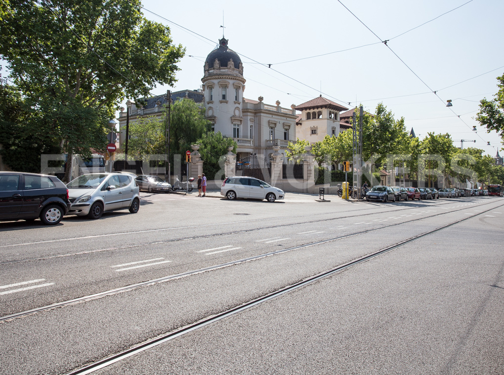 A street view of a historic mansion with a dome, surrounded by trees, parked cars, and tram tracks on a sunny day.