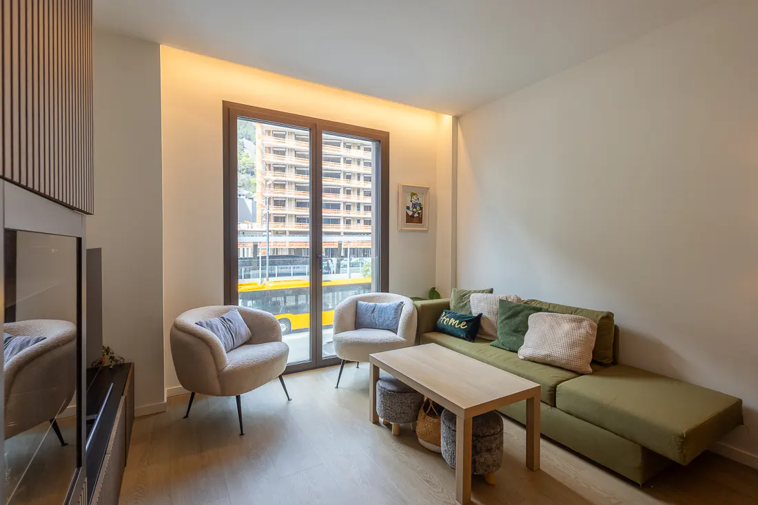 Living room with a green sofa, two chairs, and a wooden table. A large window shows a city view with a yellow bus.