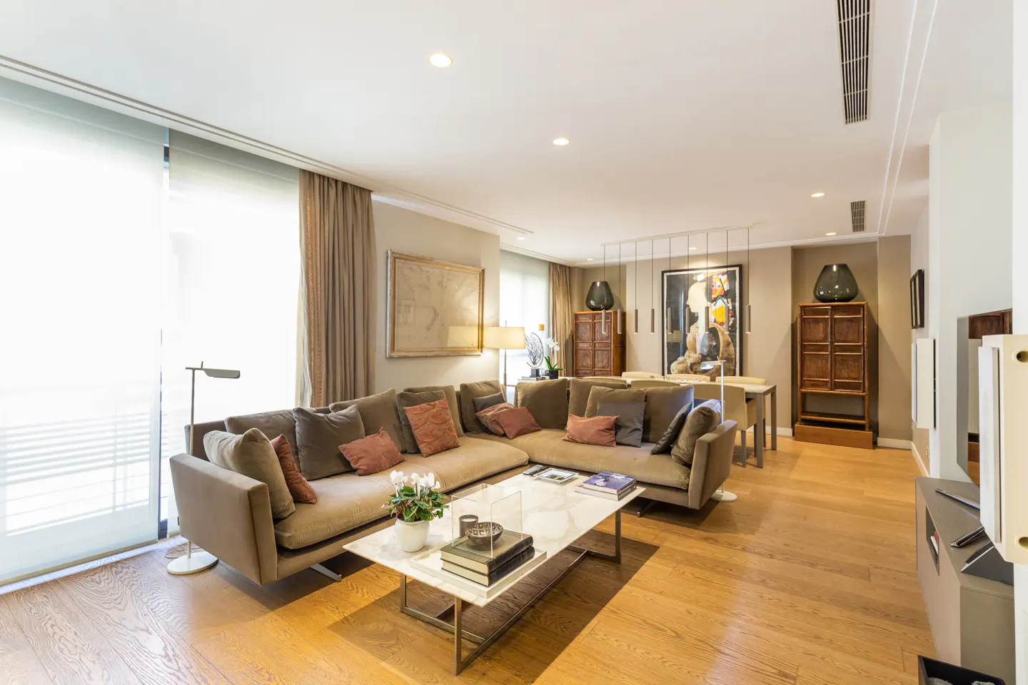 Bright living room with a brown sectional sofa, marble coffee table, and hardwood floors. Artwork and cabinets decorate the background.