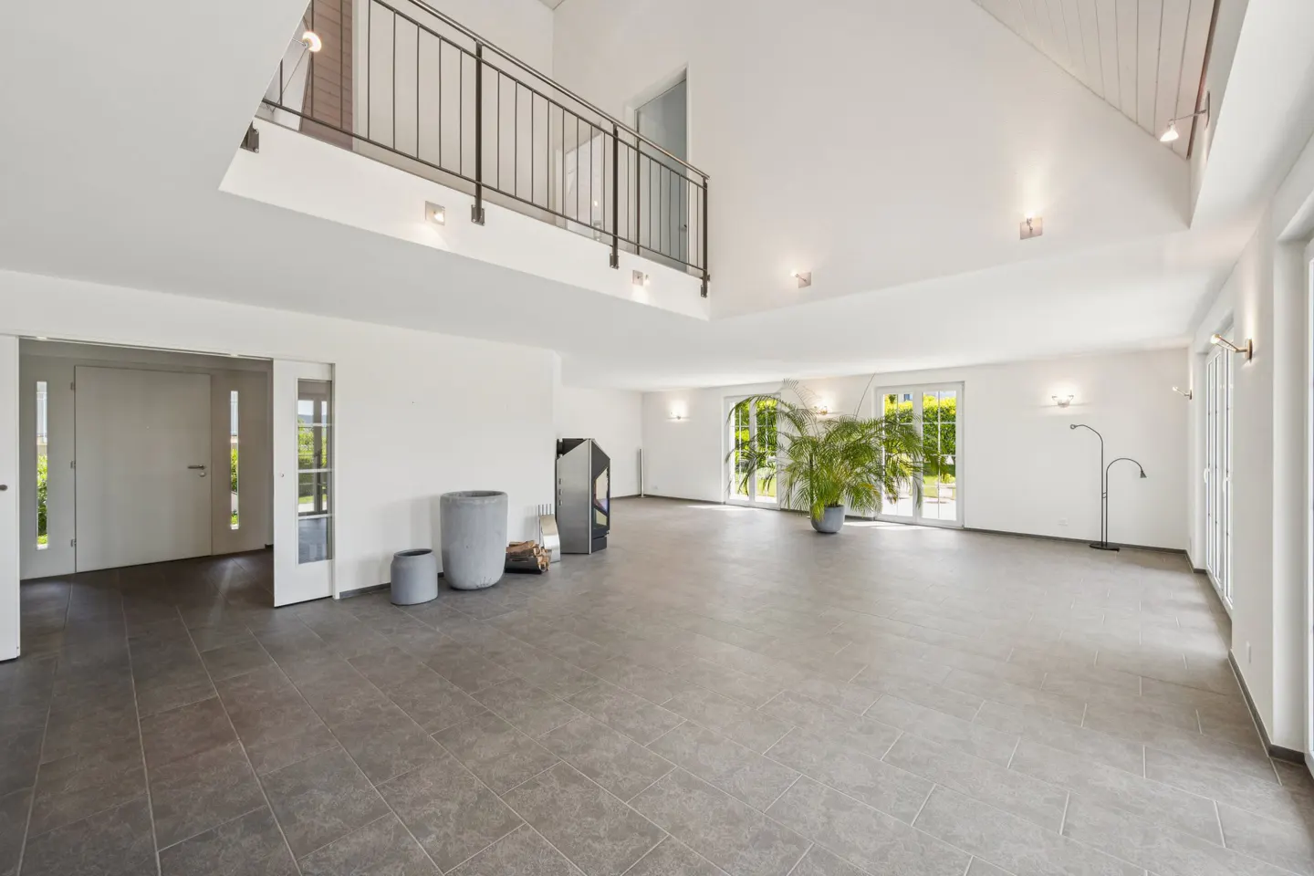 Bright, modern living room with gray tile floor, white walls, and a second-floor balcony. Large windows and a potted plant add natural light.