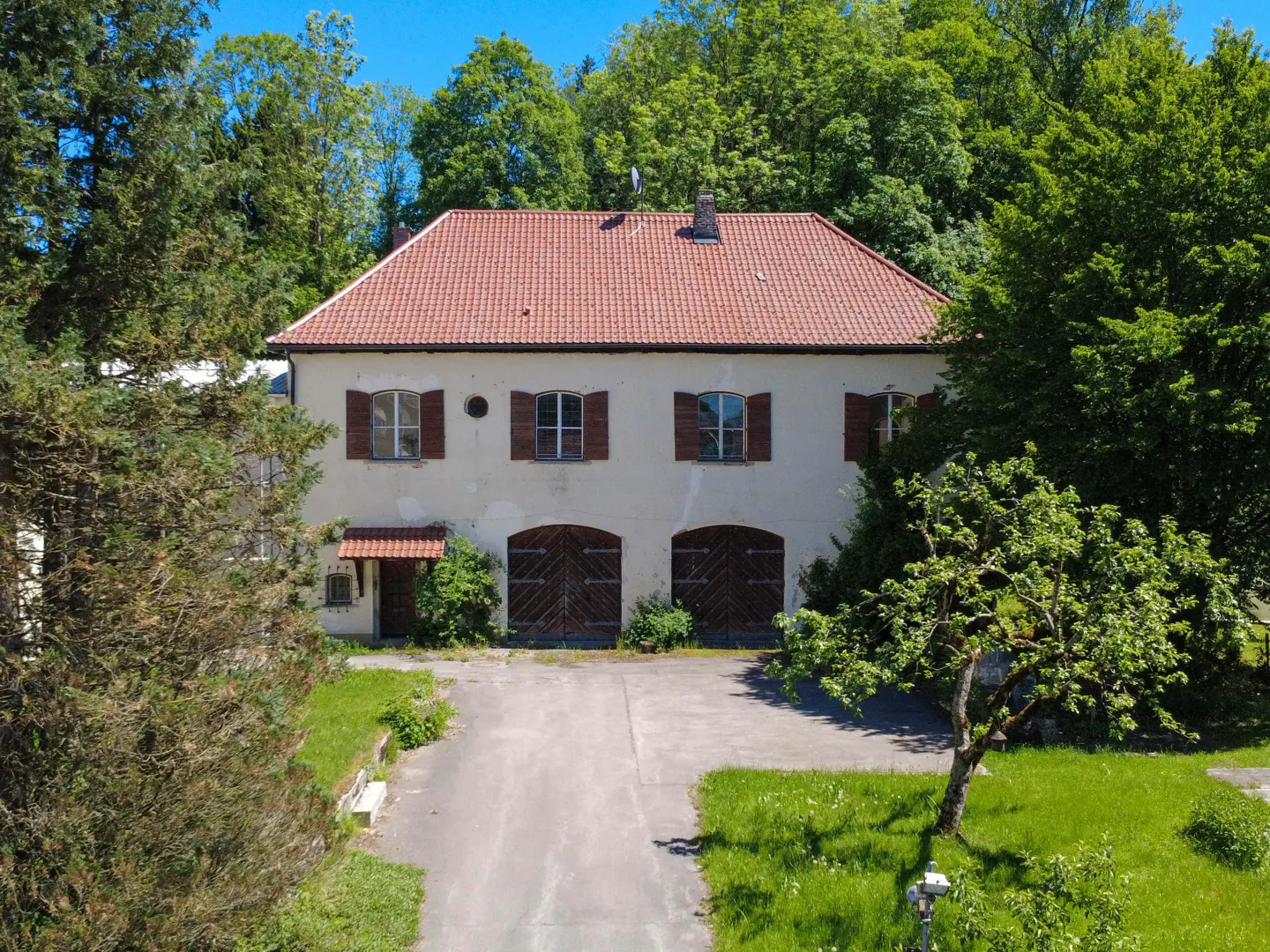 Two-story cream-colored house with a red tile roof, arched wooden garage doors, and lush green trees in the background.