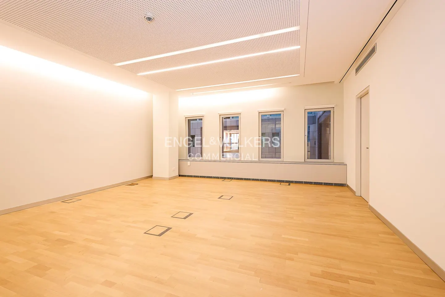 Bright, empty office space with light wood floors, white walls, and three windows. Recessed floor outlets and modern lighting.