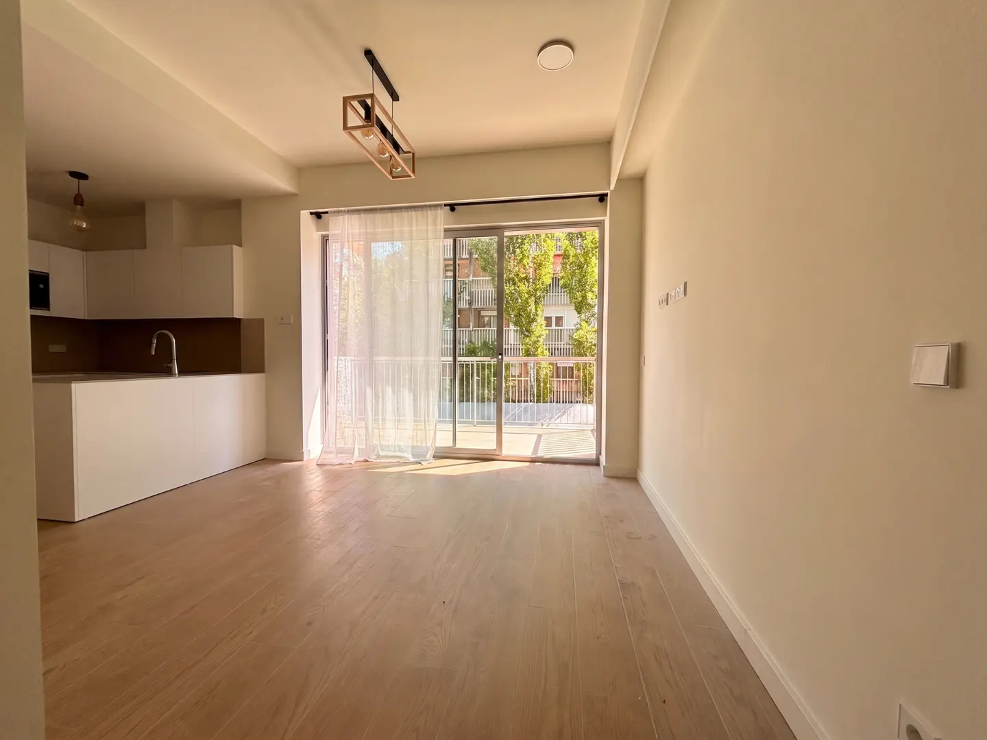 Bright, empty apartment with light wood floors, white walls, and an open kitchen. Sliding glass doors lead to a balcony with a view of trees and a building.