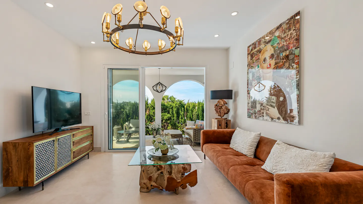 Bright living room with a brown sofa, glass coffee table with a wood base, and a large art piece on the wall. Sliding glass doors lead to a patio.