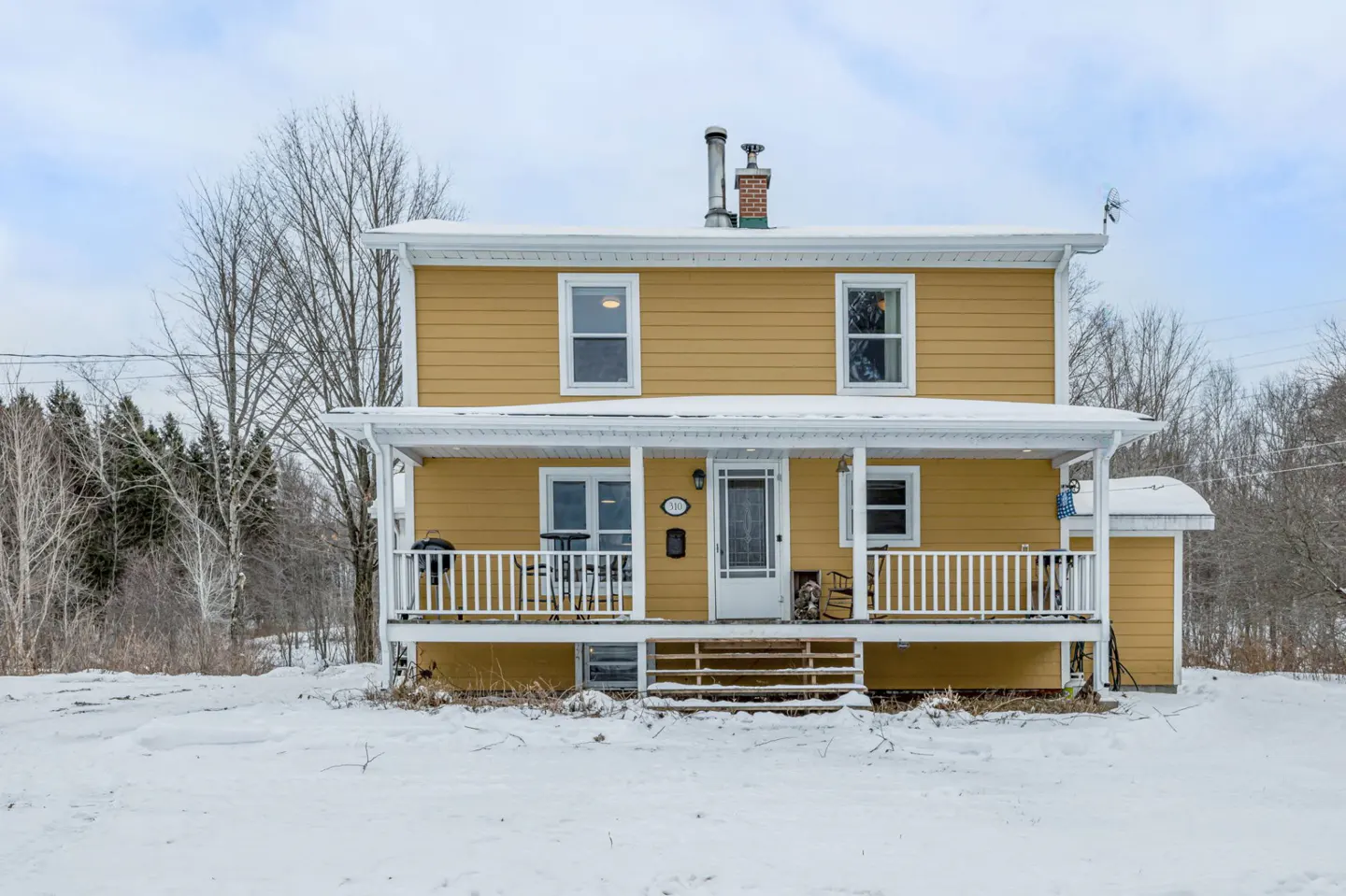 Two-story yellow house with a white porch and railing, surrounded by snow and bare trees.