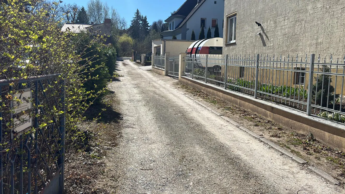 Gravel driveway leading to a house with a silver fence. Bushes and trees line the drive. A red and white trailer is parked near the house.