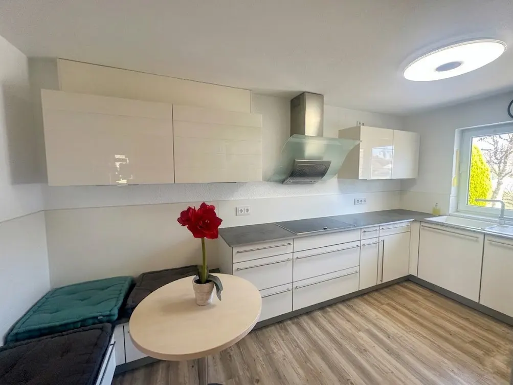 Bright kitchen with white cabinets, gray countertops, and wood-look floors. A round table with a red flower sits near a cushioned bench.