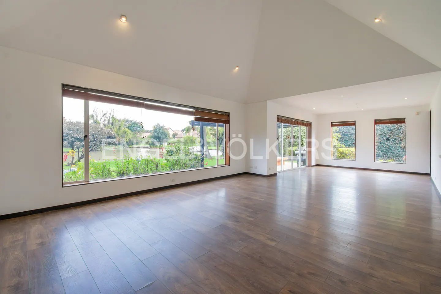 Bright, empty room with dark wood floors, white walls, and large windows overlooking a lake and greenery.