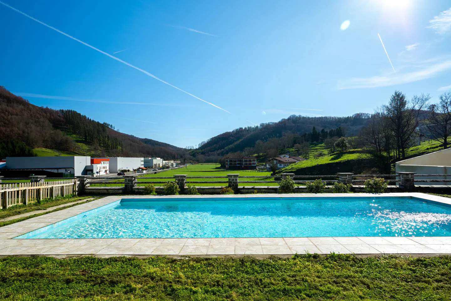 A rectangular swimming pool with sparkling blue water overlooks a green valley, distant mountains, and a clear blue sky.