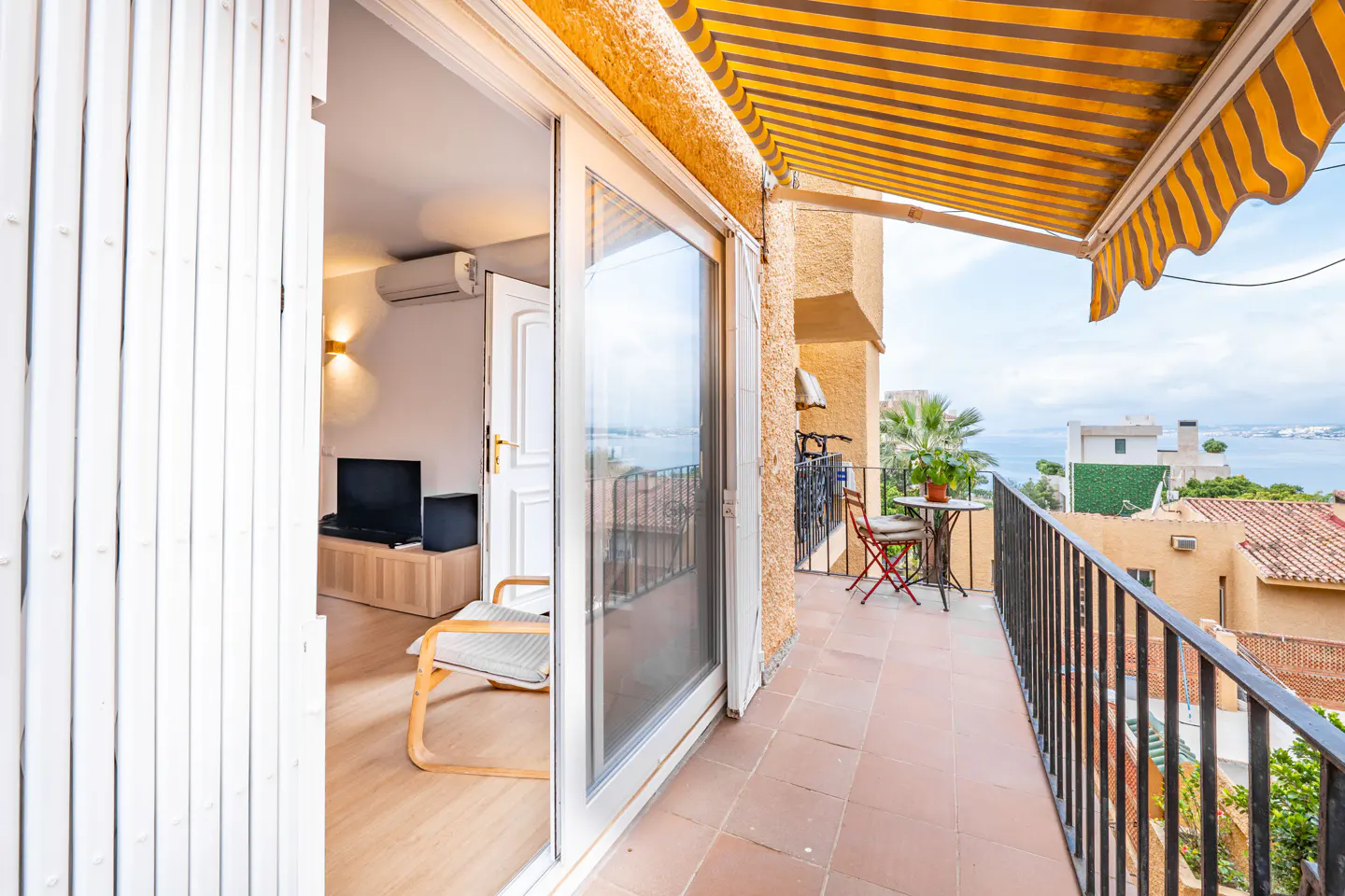 Balcony view: Open sliding glass doors reveal a living room with a chair, TV, and AC. The balcony has a table, chairs, and a yellow-striped awning.