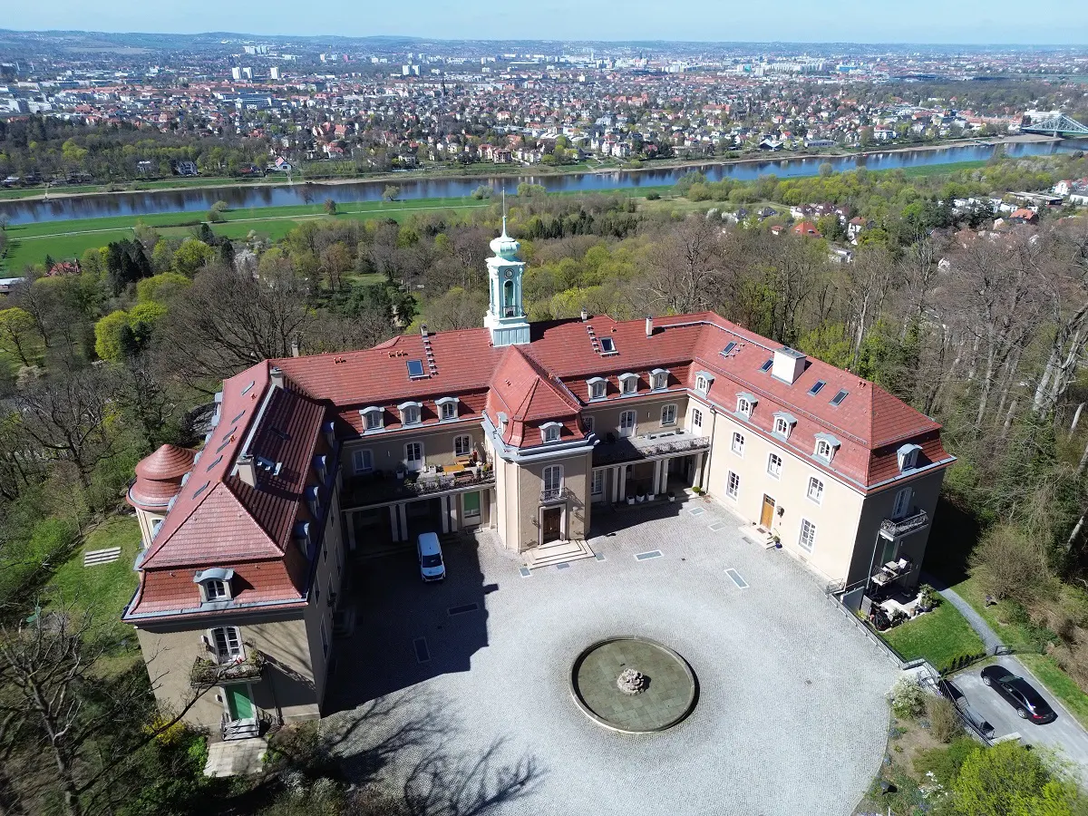 Aerial view of a large, beige building with a red tile roof and a central courtyard with a fountain. A river and city are in the background.